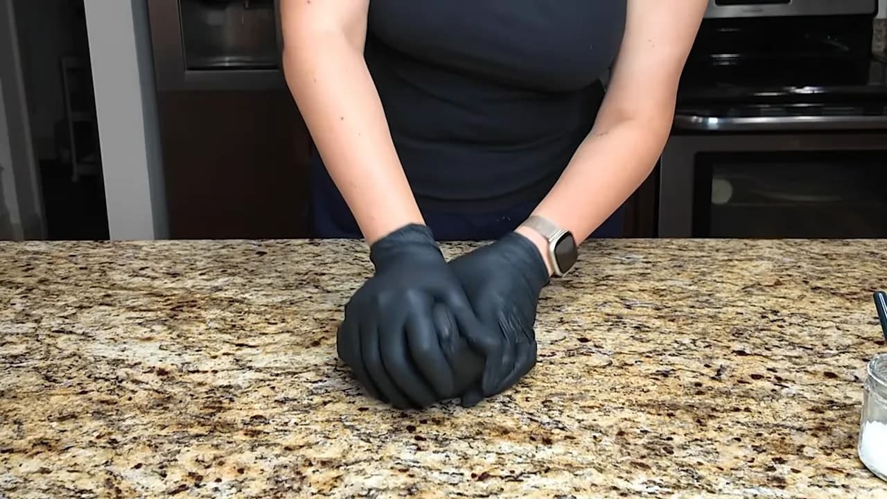 Gloved hands flattening black fondant into a smooth base on a granite countertop
